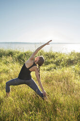 Woman practicing extended side angle pose yoga on grassy field by sea during sunny day - CAVF31297