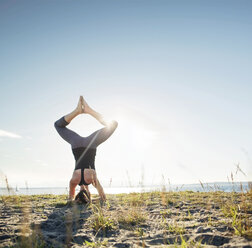 Rear view of woman practicing bound angle headstand pose on sand by sea - CAVF31300