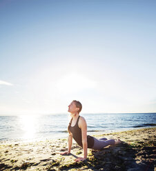 Woman practicing cobra pose at beach against sky during sunny day - CAVF31304