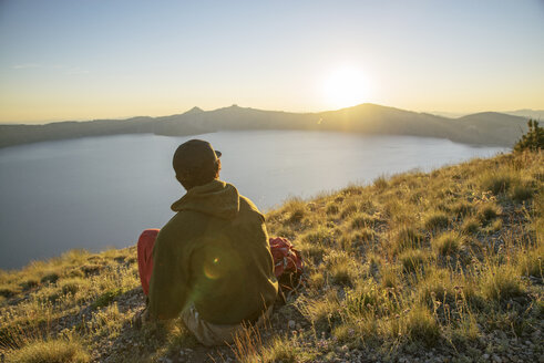 Rückansicht eines Wanderers, der während des Sonnenuntergangs auf einem Berg sitzt und die Aussicht betrachtet - CAVF31364