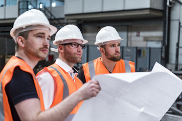 Drei Männer mit Schutzhelmen und Sicherheitswesten halten einen Plan in einer Fabrik - DIGF03579