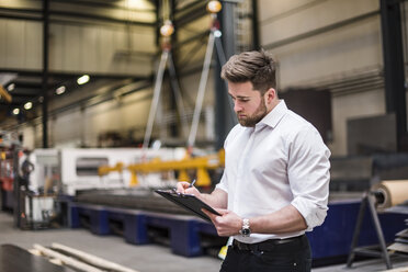 Man writing on clipboard on factory shop floor - DIGF03598