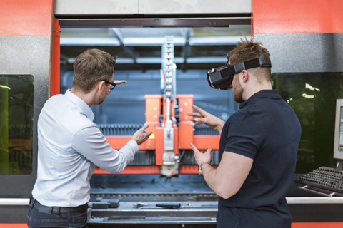 Mann erklärt einem Kollegen mit VR-Brille in einer Fabrik eine Maschine, lizenzfreies Stockfoto