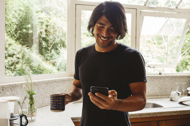 Lächelnder Mann, der in der Küche stehend ein Smartphone benutzt - CAVF31372