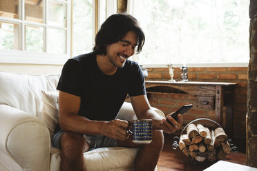 Smiling man using smart phone while sitting on armchair at home - CAVF31375