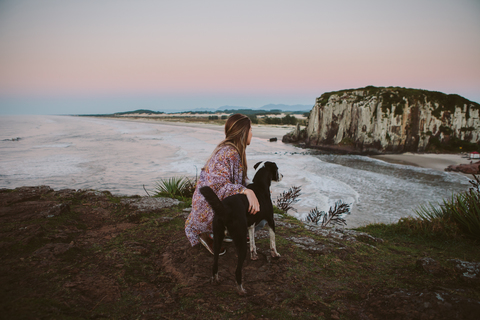 Seitenansicht einer Frau, die mit ihrem Hund auf einem Hügel am Strand kauert, während die Sonne untergeht, lizenzfreies Stockfoto