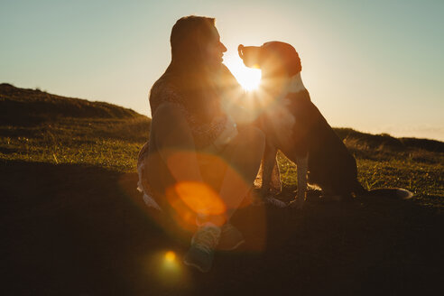Frau und Hund schauen sich an, während sie bei Sonnenuntergang auf einem Hügel sitzen - CAVF31390