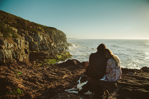 Paar und Hund sitzen auf einem Felsen am Meer gegen den klaren Himmel, lizenzfreies Stockfoto