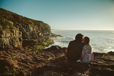 Rückansicht eines Paares, das sich küsst, während es auf einem Felsen am Meer sitzt, gegen einen klaren Himmel - CAVF31392