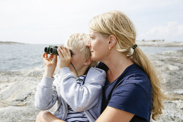 Mutter und Sohn sitzen am felsigen Meeresufer, der Sohn schaut durch ein Fernglas in den Stockholmer Schären - FOLF05734