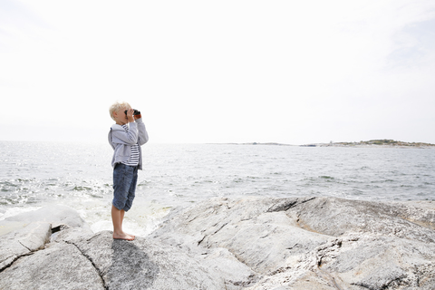 Junge steht am felsigen Meeresufer und schaut durch ein Fernglas in den Stockholmer Schären, lizenzfreies Stockfoto