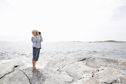 Boy standing on rocky seashore and looking through binoculars in the Stockholm archipelago - FOLF05736