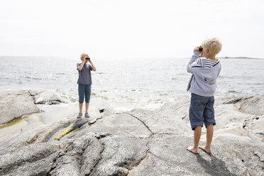 Two boys standing on rocky seashore and looking through binoculars in the Stockholm archipelago - FOLF05738