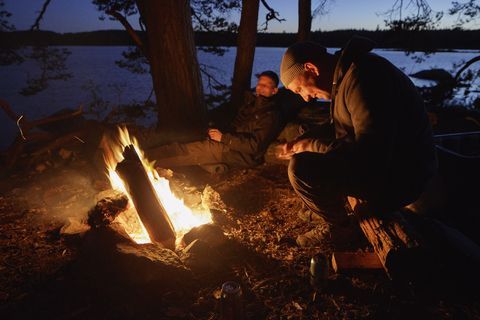 Mittlere erwachsene Männer sitzen in der Abenddämmerung am Lagerfeuer, lizenzfreies Stockfoto