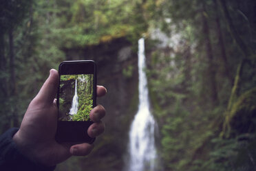Cropped image of man photographing waterfall through smart phone in forest - CAVF31691