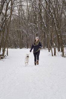 Glückliche Frau, die mit ihrem Hund auf einem schneebedeckten Feld vor kahlen Bäumen spazieren geht - CAVF31700