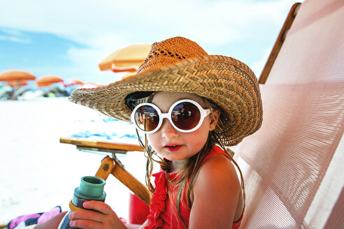 Porträt eines Mädchens mit Sonnenbrille und Strohhut, das auf einem Liegestuhl am Strand sitzt - CAVF31708