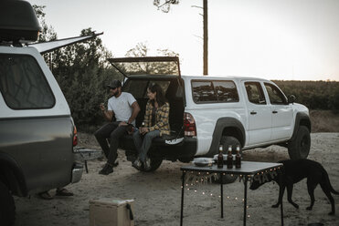 Paar sitzt im Kofferraum eines Autos auf dem Campingplatz mit Hund gegen den klaren Himmel - CAVF31739