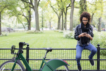 Woman with bicycle using mobile phone while sitting on railing in park - CAVF31906