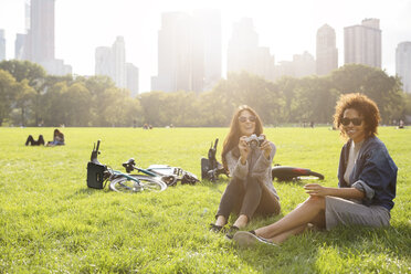 Happy woman holding camera while relaxing with friend on grassy field - CAVF31917