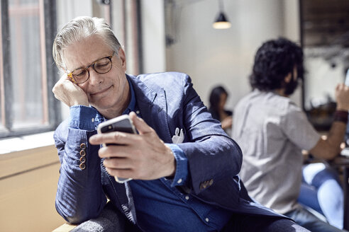 Geschäftsmann schaut auf sein Smartphone, im Hintergrund sitzt ein männlicher Kollege - CAVF32003