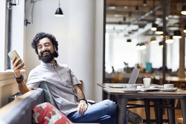 Happy businessman looking at smart phone while sitting in office cafeteria - CAVF32020