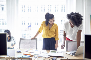 Businesswomen pointing towards document while standing against window with female colleague sitting in background - CAVF32044