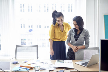 Happy businesswomen looking at documents while standing against window in office - CAVF32045