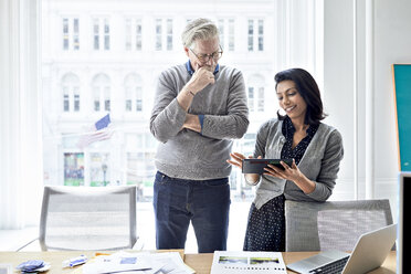 Businesswoman showing tablet computer to senior businessman while standing against window in creative office - CAVF32048