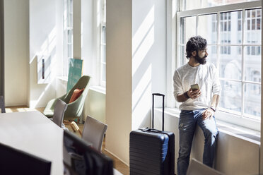 Geschäftsmann schaut durch ein Fenster und hält ein Smartphone in einem kreativen Büro - CAVF32064