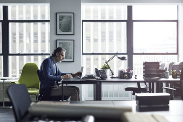 Geschäftsmann mit Laptop bei der Arbeit gegen das Fenster im Büro - CAVF32067