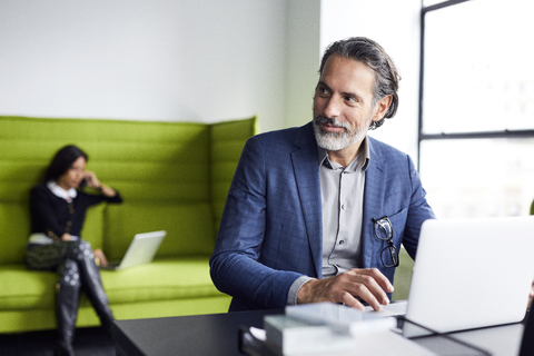 Geschäftsmann schaut weg, während er einen Laptop benutzt und eine Kollegin im Hintergrund arbeitet, lizenzfreies Stockfoto