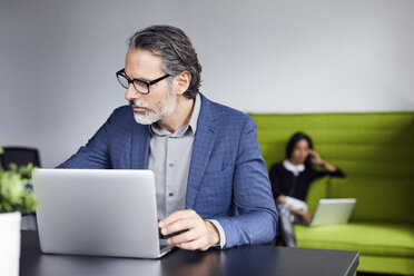 Businessman looking away while using laptop computer with colleague working in background - CAVF32073