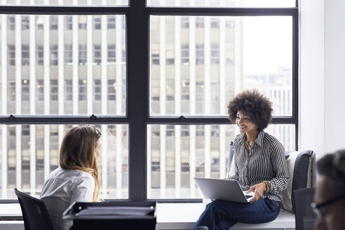 Happy businesswoman discussing with female colleague while sitting on window sill in office - CAVF32085