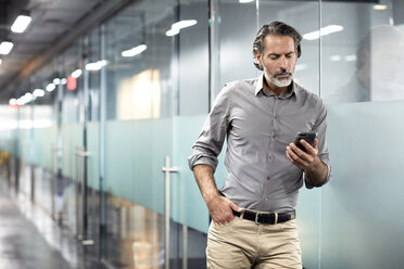 Geschäftsmann mit Hand in der Tasche, der auf sein Smartphone schaut, während er in der Büro-Lobby steht - CAVF32107