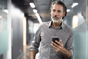 Businessman looking away while holding smart phone in office lobby - CAVF32108