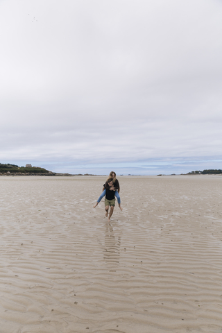Frankreich, Bretagne, Guisseny, junger Mann läuft mit Freundin huckepack am Strand, lizenzfreies Stockfoto