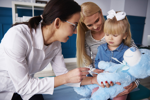 Arzt und Mädchen mit Mutter beim Impfen eines Teddys in einer Arztpraxis, lizenzfreies Stockfoto