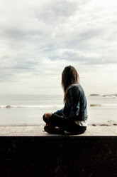 Junge Frau sitzt auf einer Mauer und schaut auf das Meer - MGOF03747