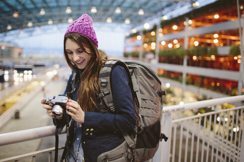 Lächelnde Frau mit Rucksack, die in die Kamera schaut, während sie sich auf ein Geländer im Flughafen stützt - CAVF32122