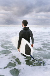 Rückansicht eines Mannes, der ein Surfbrett trägt, während er im Meer gegen einen bewölkten Himmel läuft - CAVF32155