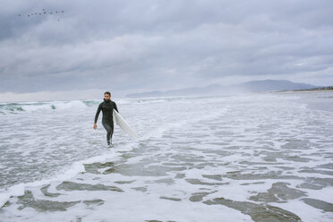 Mann mit Surfbrett schaut weg, während er im Meer gegen den bewölkten Himmel läuft - CAVF32158