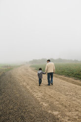 Rückansicht von Vater und Sohn, die auf einem Feldweg gegen den Himmel bei nebligem Wetter laufen - CAVF32168