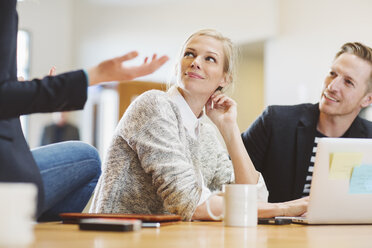 Colleagues looking at businesswoman talking while sitting on table in office - CAVF32222