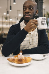 Mann mit Croissant und Tasse Kaffee in einem Café, der sich umschaut - MAUF01359