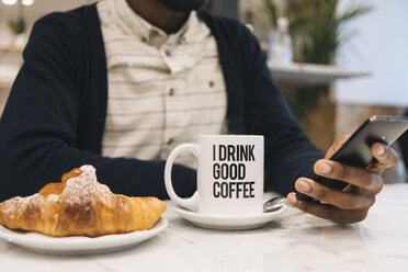 Nahaufnahme eines Mannes mit Croissant und Tasse Kaffee in einem Café, der sein Handy benutzt - MAUF01360