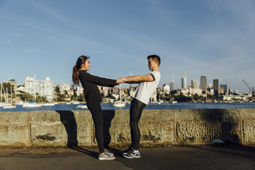 Young couple holding hands while stretching on promenade against cityscape - CAVF32324