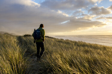 Rear view of man standing on grassy field by sea against sky during sunset - CAVF32347