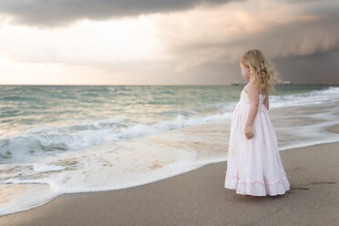 Seitenansicht eines Mädchens in einem Kleid, das am Strand vor einem bewölkten Himmel bei Sonnenuntergang steht - CAVF32359