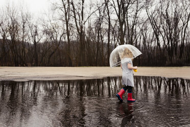 Seitenansicht eines Mädchens, das einen Regenschirm trägt, während es in der Regenzeit auf einer Pfütze läuft - CAVF32361